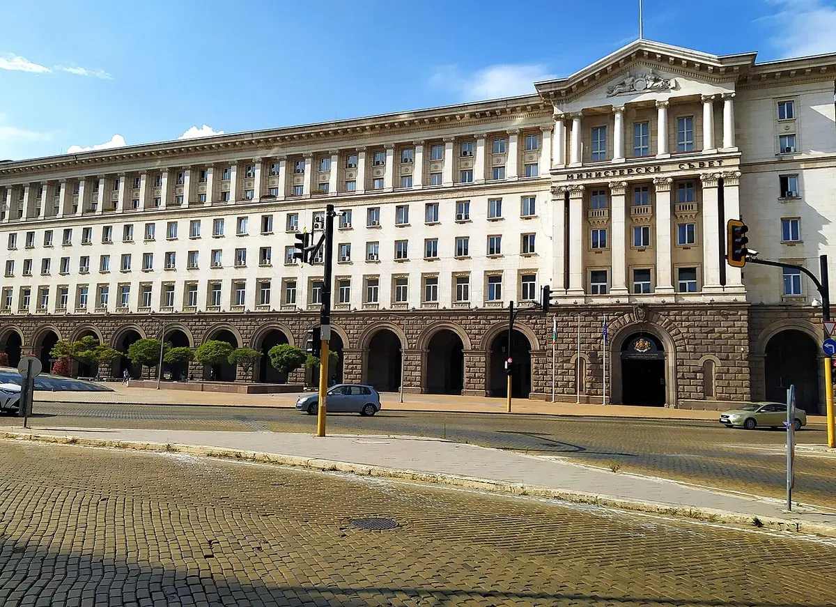 Сградата на Министерския съвет в София с надпис над колоните, жълти павета и автомобили отпред